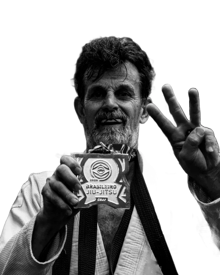 Camilo Scuzziato sorrindo, de kimono branco, mostrando a medalha do Brasileiro de Jiu-Jitsu 2026 e fazendo o número três com a outra mão